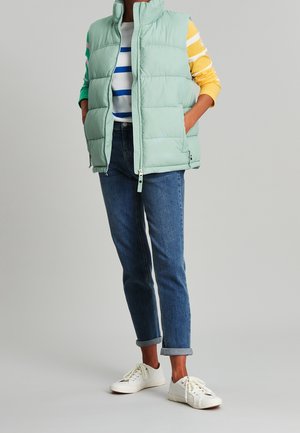 Person iført en lysegrøn dunvest, stribet langærmet skjorte, blå opkøbte jeans og hvide sneakers, stående med hænderne i vestens lommer.