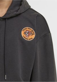 Sötétszürke kapucnis pulóver kenguruzsebbel, narancssárga kör alakú folttal, amelyen egy tigris és a "TRUE RELIGION TIGERS MFG" felirat látható.