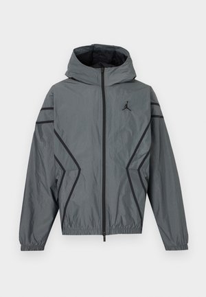 Sivá vetrovka s kapucňou, predný zips, čierne akcentové čiary, elastické manžety, bočné vrecká a malé čierne logo na hrudi.