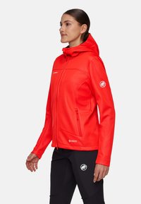 Frau trägt hellrote Softshelljacke mit Kapuze und schwarze Hose mit weißen Mammut-Logos, steht im Profil vor weißem Hintergrund.
