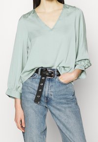 Minzgrünes V-Ausschnitt-Bluse mit langen, gepufften Ärmeln, kombiniert mit hoch taillierten blauen Jeans und einem schwarzen Gürtel mit metallischen Beschlägen.