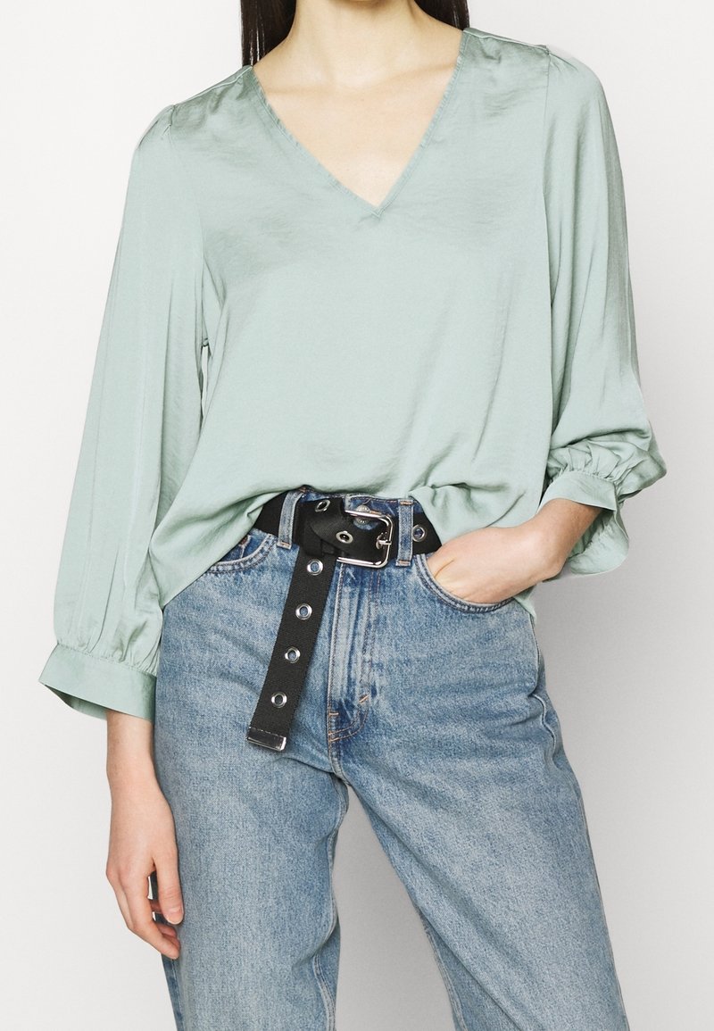 Minzgrünes V-Ausschnitt-Bluse mit langen, gepufften Ärmeln, kombiniert mit hoch taillierten blauen Jeans und einem schwarzen Gürtel mit metallischen Beschlägen.