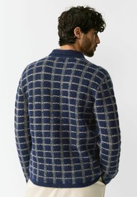 Tmavomodrý pletený sveter s mriežkovým vzorom v krémovej farbe. Textúra je mäkká, so zaobleným golierom a dlhými rukávmi.