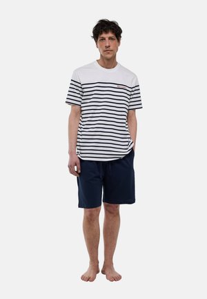 Homme debout pieds nus portant une chemise à manches courtes blanche et bleu marine à rayures, ainsi qu'un short uni bleu marine arrivant aux genoux sur un fond uni.