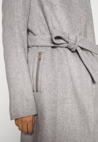 Cappotto di lana grigio chiaro con vita regolabile annodata e tasca verticale con zip sul lato sinistro, indossato da una persona.