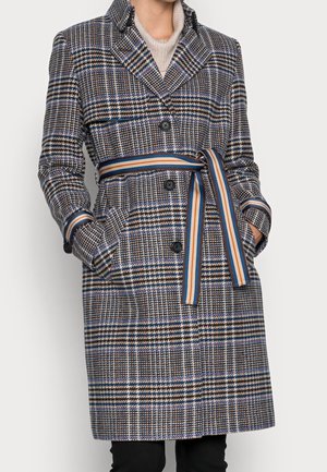 Frau trägt einen knieumspielenden Hahnentritt-Mantel mit schwarzen Knöpfen und einem gestreiften Gürtel, der in der Taille gebunden ist, Hände in den Taschen.