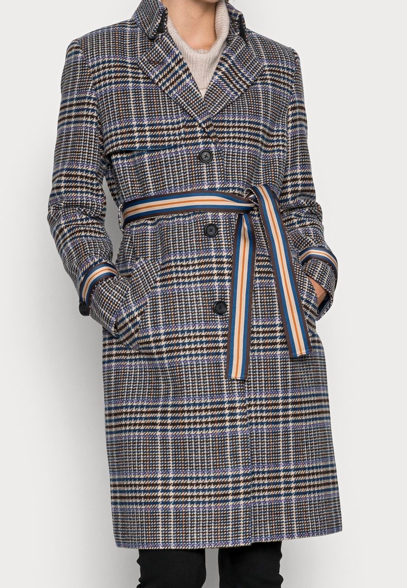 Femme portant un manteau mi-long à motif pied-de-poule avec des boutons noirs et une ceinture rayée nouée à la taille, les mains dans les poches.
