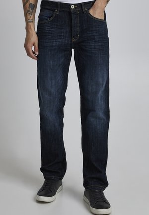 Mørkeblå denimjeans med rette ben, med subtil falming, gule sømmer og frontlommer. Brukt med mørke sneakers.