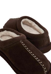Un paio di pantofole marroni in suede con rivestimento in peluche bianco e cuciture decorative bianche lungo i bordi superiori, progettate per il comfort interno.