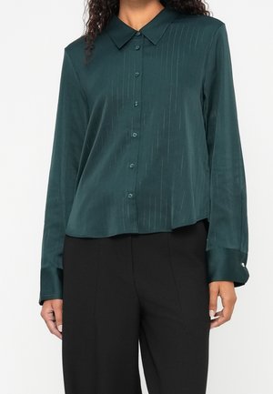 Kobieta w ciemnozielonej koszuli na guziki z subtelnymi pionowymi paskami i czarnych spodniach, pokazana od szyi do bioder na jednolitym tle.