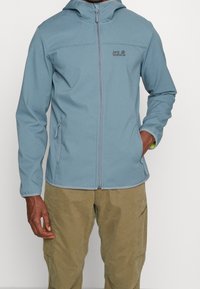 Giacca softshell blu chiaro con zip frontale, cappuccio regolabile e tasche laterali. Presenta un logo sul petto e un tessuto resistente.