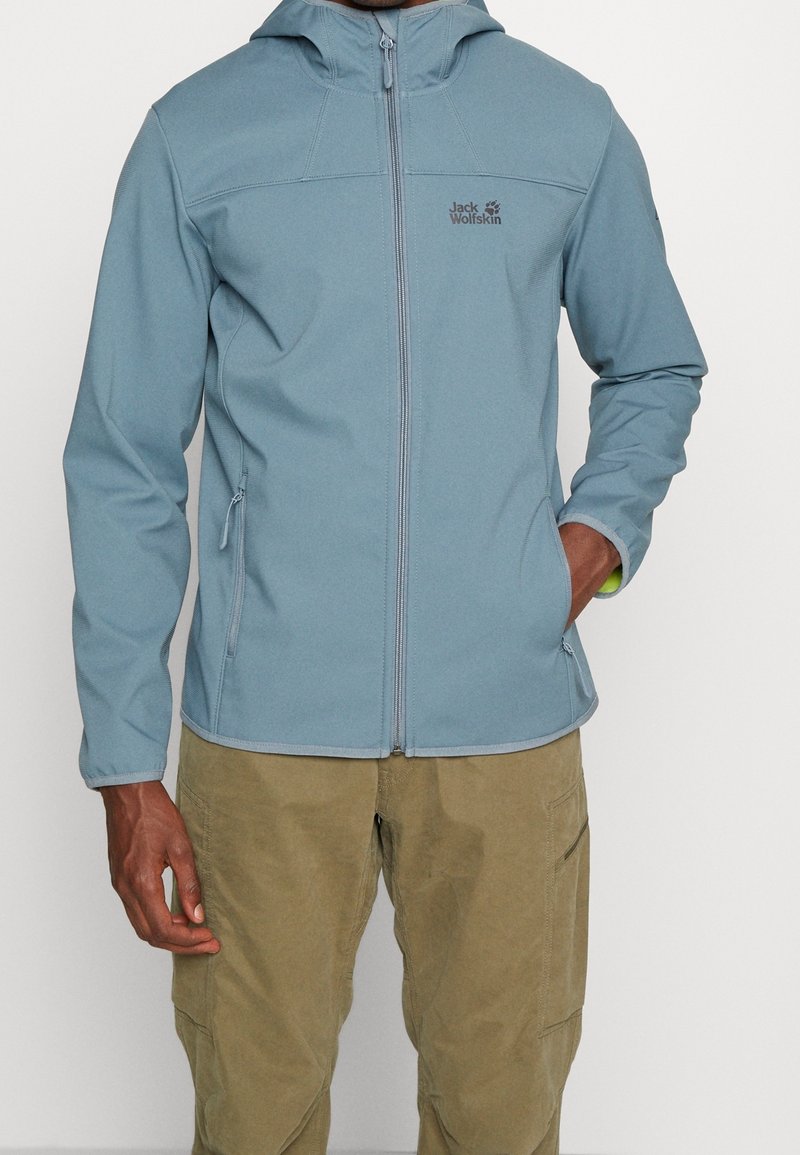 Giacca softshell blu chiaro con zip frontale, cappuccio regolabile e tasche laterali. Presenta un logo sul petto e un tessuto resistente.