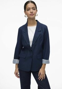 Tumesinine bleiser, millel on kohandatud lõige, kaks esist taskut ja triibulised varruka detailid. Kombineeritud valge särk ja tumedad teksapüksid.