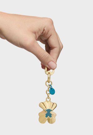 Ręka trzymająca złoty brelok z niebieskim wycięciem w kształcie niedźwiadka i małą niebieską okrągłą zawieszką na białym tle.