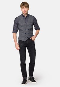 Szary w kratkę kamizelka na ciemnej koszuli, zestawiona z czarnymi przetartymi dżinsami i czarnymi sneakersami. Kamizelka posiada podwójne guziki.