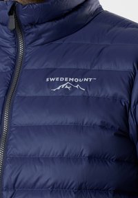 Marineblaues gepolstertes Westchen mit hohem Kragen, Reißverschluss vorne und horizontalem Steppmuster. Mit einem weißen Logo auf der Brust.