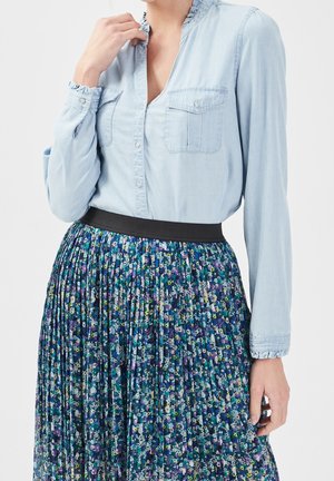 Femme portant une chemise bleu clair à boutons avec col et poignets à volants, associée à une jupe plissée fleurie avec ceinture noire.