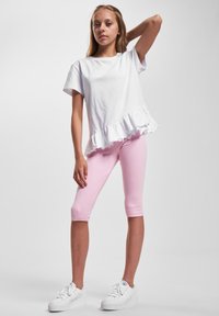 Hvit bomulls T-skjorte med volangkant, kombinert med tettsittende rosa leggings. Modellen har på seg hvite sneakers med en glatt tekstur gjennom hele.