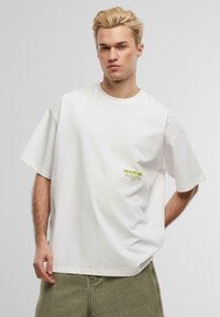 White cotton t-shirt with oversized fit, round neck, and green logo printed on the left side. Smooth texture, short sleeves.