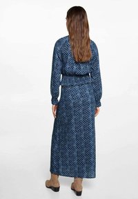 Maksi obleka iz temno modrega blaga z geometrijskim vzorcem. Dolgi rokavi, elastičen pas in zračna krila. Obuta z bež obutvijo.