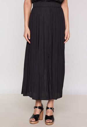 Jupe maxi plissée noire avec une taille haute et une texture fluide, présentée avec des sandales à talons hauts noires à lanières.