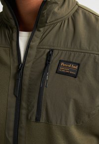Olijfgroene, ritsjack met textuurstof, voorzien van een borstzak, zwarte rits en oranje logopatch. Lichtgewicht en functioneel ontwerp.