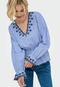 Blouse rayée bleue et blanche avec un décolleté en V, taille élastique et manches évasées présentant une broderie florale le long de l'encolure et des poignets.