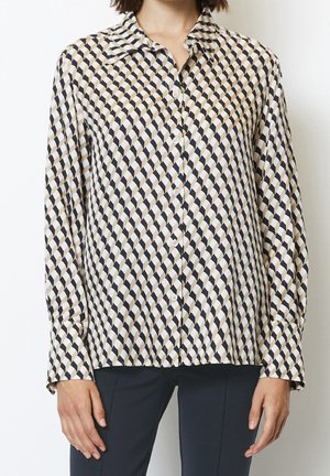 Personne portant une chemise à manches longues boutonnée avec un motif à carreaux diagonaux en beige, noir et blanc, associée à un pantalon foncé.