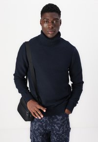 Marineblå strikket genser med høy hals og ribbestrikket krage og mansjetter, kombinert med mørke mønstrede cargo-bukser og en crossover-veske.