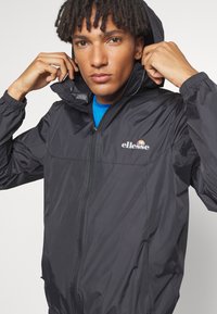 Schwarze leichte Jacke mit einem Reißverschluss und Kapuze. Verfügt über elastische Bündchen, Seitentaschen und ein kleines Logo auf der Brust.