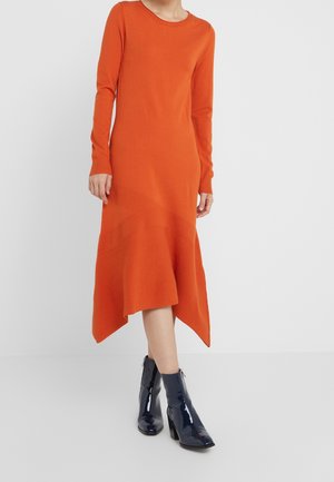 Frau trägt ein langärmeliges, asymmetrisches Midikleid in Orange mit diagonalem Rippmuster, kombiniert mit glänzenden schwarzen Stiefeletten.
