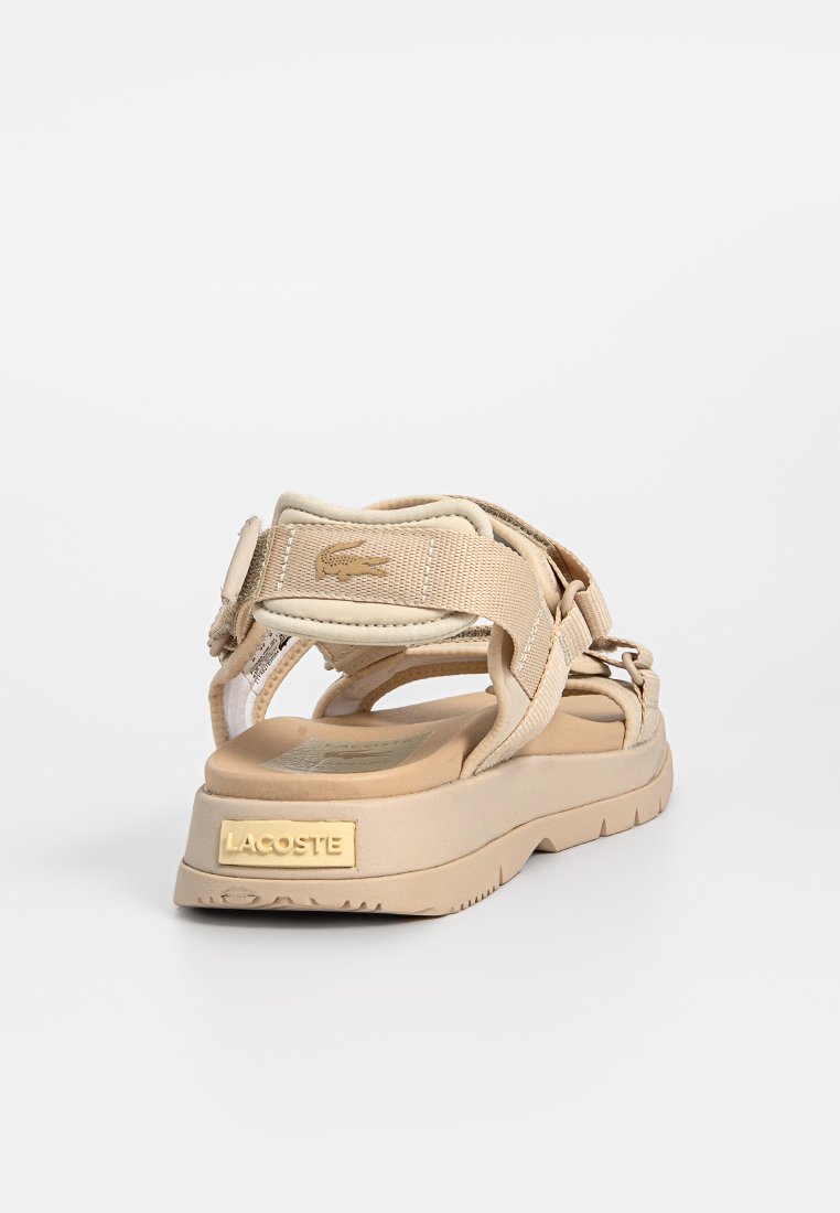 Sandale beige avec des sangles en tissu réglables, semelle texturée, semelle rembourrée et logo Lacoste embossé sur le talon. Design léger et moderne.