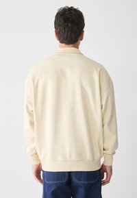 Sudadera de color beige claro con un ajuste relajado, hombros caídos y puños acanalados. Textura suave y diseño simple en la parte posterior. Jeans visibles por debajo.
