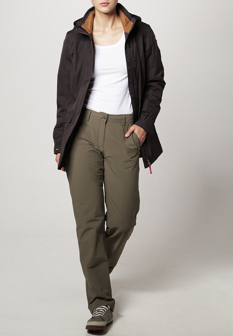 Personne portant une veste à capuche noire, un top blanc sans manches, un pantalon vert olive et des baskets grises, marchant avec une main dans la poche.