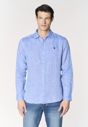 Uomo che indossa una camicia di lino azzurro chiaro con bottoni e maniche lunghe e jeans blu, in piedi davanti a uno sfondo bianco uniforme.