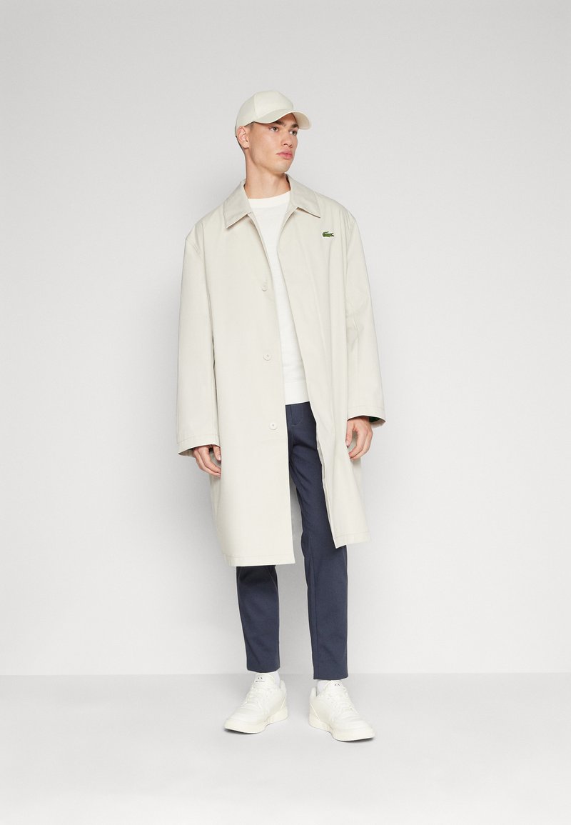 Cappotto lungo beige chiaro con colletto a rever, chiusura a bottoni e accento con logo verde. Indossato sopra un maglione bianco, pantaloni blu scuro e sneakers bianche.