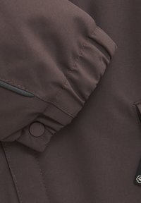 Brun jacka av tyg med strukturerad yta, med rynkade ärmslut, en tryckknapp och en liten logotagg på sidan.