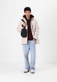 Puffjacke in hellem Beige mit Kapuze, Frontreißverschluss und schwarzen Akzenten. Dunkelbrauner Pullover darunter und blaue Jeans. Schwarze Umhängetasche.
