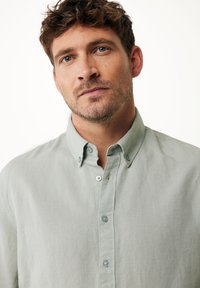 Světle zelená košile na knoflíky s límečkem, s jemnou strukturou a decentním leskem, zapínaná modrými knoflíky.