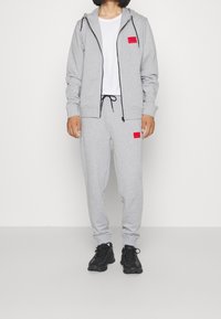 Conjunto de sudadera y pantalones deportivos grises hechos de algodón. Chaqueta con capucha y detalles negros, con parche de logo rojo. Zapatillas negras con suelas texturizadas.