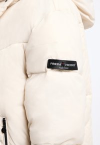 Kremowa kurtka puchowa z błyszczącego materiału, z czarną metką z logo "FRIEDA FREDDIES" umieszczoną na lewym rękawie.