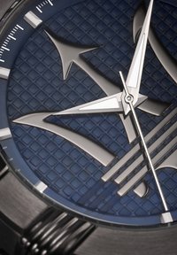 Metalen horloge met een blauwe gestructureerde wijzerplaat, zilveren wijzers en gedurfde logo-details. Kenmerkt een gemustere achtergrond en glanzende accenten.