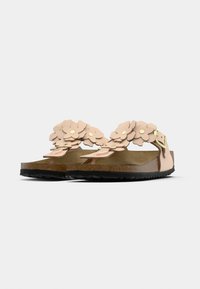 Μπεζ σανδάλια με φελοειδείς σόλες, μαύρη σόλα με ανάγλυφο σχέδιο και στρωματοποιημένες διακοσμήσεις  λουλουδιών στην κορυφή, στερεωμένα με λουράκι και χρυσή αγκράφα.