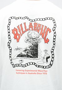 Λευκό βαμβακερό μπλουζάκι με γραφικό σχέδιο. Περιλαμβάνει τη λέξη «BILLABONG» με κόκκινα γράμματα, συνοδευόμενη από απεικονίσεις νερού, σέρφερ και ενός χαρακτήρα.