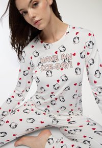 Pigiama a maniche lunghe bianco con motivi di pinguini e cuori. Presenta una grafica con la scritta "PASSA UNA GIORNATA FREDDA!" in rosa. Tessuto morbido e elastico.
