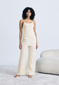 Γυναίκα με σγουρά μαλλιά στέκεται ξυπόλητη σε χαλί, φοράει ανοιχτό κίτρινο αμάνικο τοπ και παντελόνια με πλατύ πόδι που ταιριάζουν, δίπλα σε κρεμ καμπυλωτό καναπέ.