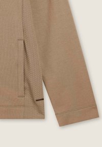 Maglia a maniche lunghe marrone con pannelli laterali testurizzati e una tasca laterale. Presenta una finitura liscia e dettagli di cucitura sottili lungo le cuciture.
