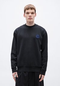 Μαύρο βαμβακερό φούτερ με μακριά μανίκια, ελαστικές μανσέτες και στρογγυλή λαιμόκοψη, με μια σκούρα μπλε κεντημένη λογότυπο στο στήθος.