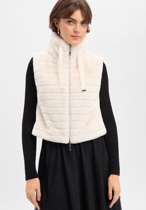 Jeune femme portant une veste sans manches en fausse fourrure blanche zippée sur une robe noire à manches longues, debout devant un fond clair uni.