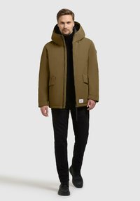 Olijfgroene gewatteerde jas met een capuchon, voorzien van een rits aan de voorkant, twee zijzakken en een contrastvoering. Draag het over een zwarte coltrui en broek.